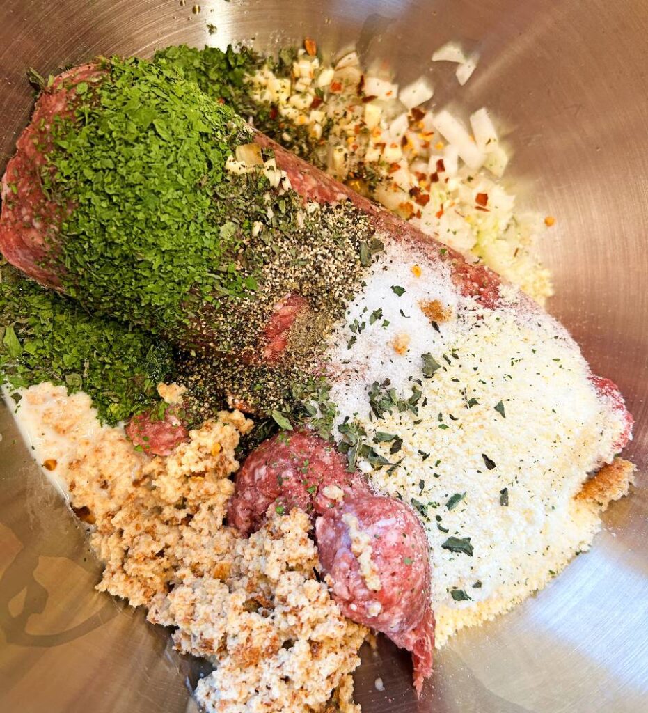 Ground beef meatball mixture in a large mixing bowl with breadcrumbs, Parmesan, onion, and herbs.