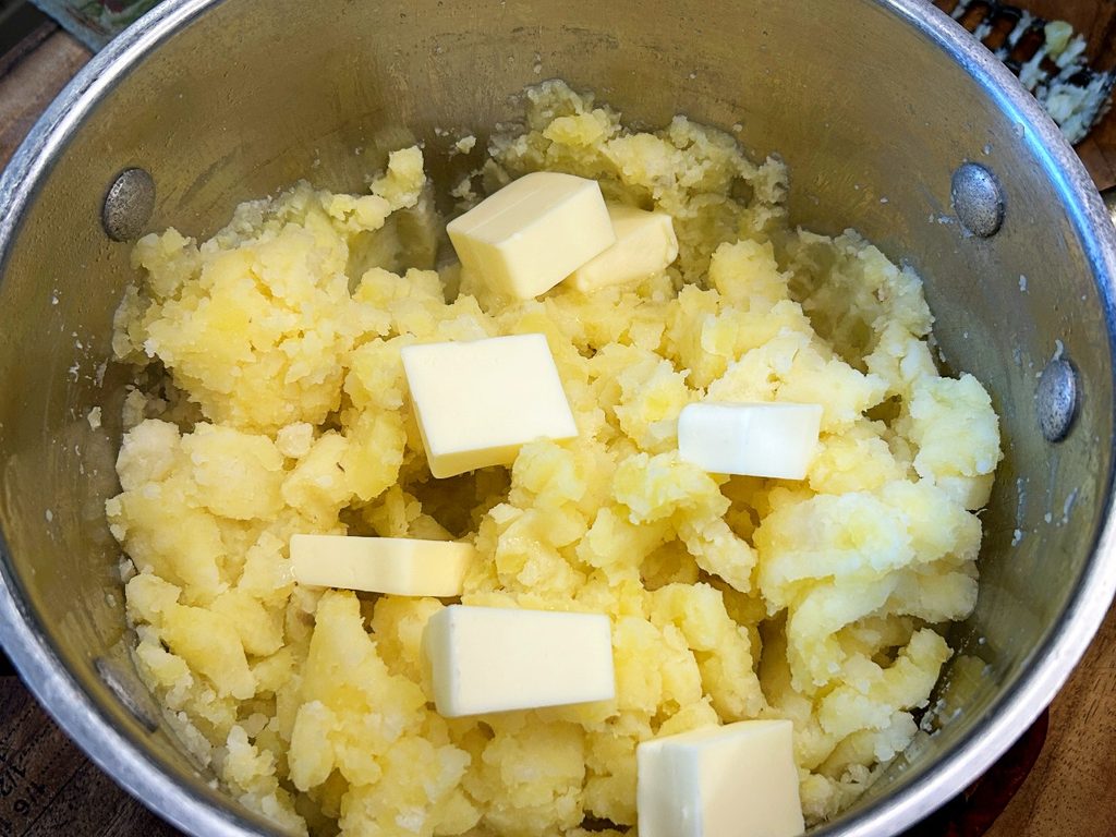 Cooked potatoes being mashed with butter for creamy mashed potatoes