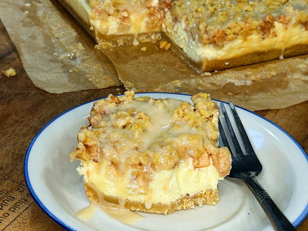 Apple cheesecake bar with cinnamon-spiced apples, crumble topping and maple glaze, with more cheesecake bars in the background.